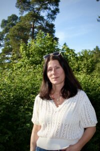 En kvinna med solglasögon på huvudet framför ett buskage i sommarsol.
