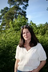 En kvinna med solglasögon på huvudet framför ett buskage i sommarsol.
